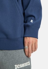 Námorníckomodrá mikina s rebrovanými manžetami, s malým bielym logom na rukáve. Sivé tepláky s bielym nápisom značky. Povrch je mäkký.