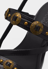 Μαύρα λουράκια σανδαλιών με στρογγυλές χρυσές καρφίτσες που απεικονίζουν αστέρια και τετράγωνες μαύρες κεντρικές λεπτομέρειες, σε κοντινή λήψη.