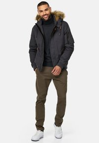 Chaqueta bomber negra para hombre con capucha forrada de piel, bolsillos con cremallera, combinada con chinos oliva y zapatillas blancas. Textura suave con un diseño casual.