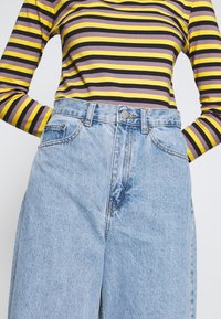 Randig långärmad topp i svart, gult och lila, matchad med högmidjade blå denimjeans som har framfickor och metallknäppning.