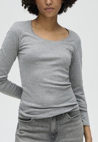 Woman wearing a grey long-sleeve shirt and grey jeans, with one hand behind her back against a plain light background.
