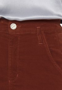 Pantalones de pana marrón con un botón metálico plateado, costuras en contraste y bolsillos laterales. Textura suave con un acabado ligeramente brillante.