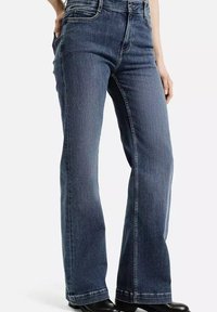 Osoba w ciemnoniebieskich dżinsach bootcut z rękami w kieszeniach, w czarnych butach, stojąca na jednolitym białym tle.