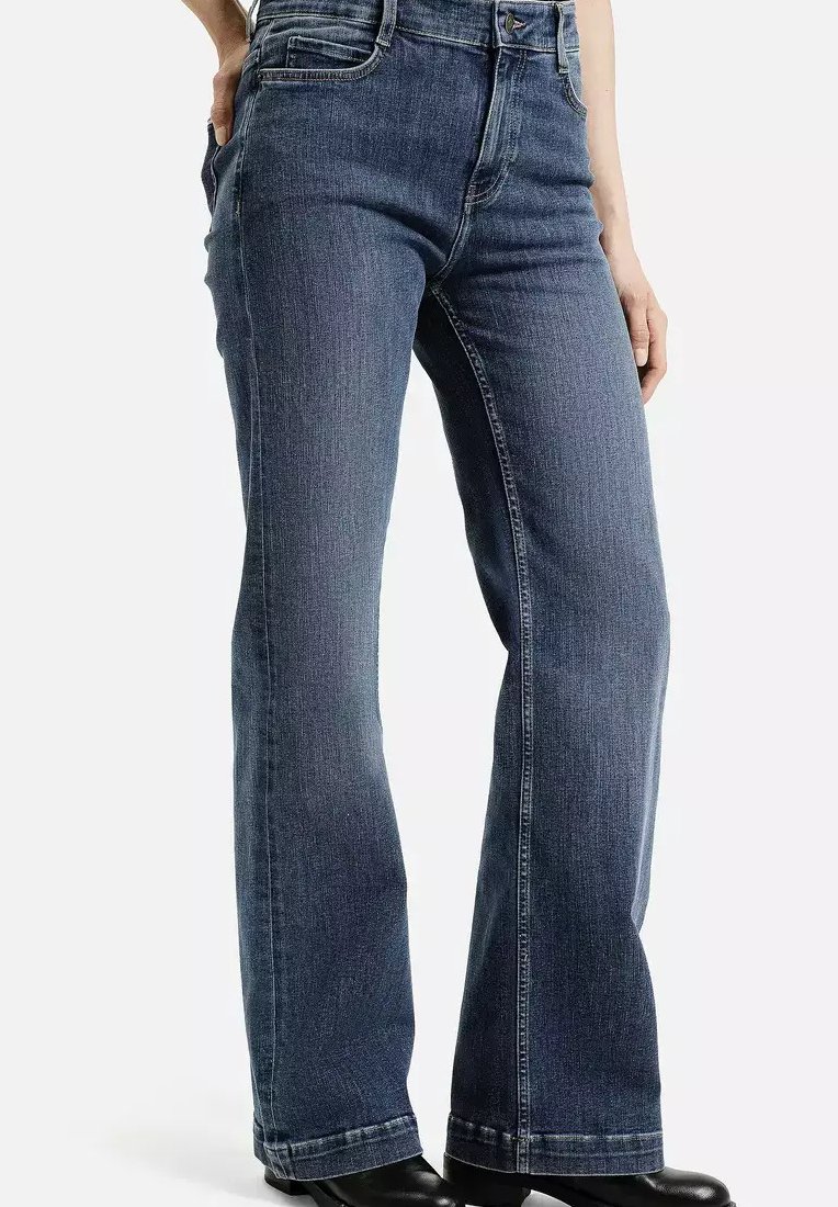 Osoba w ciemnoniebieskich dżinsach bootcut z rękami w kieszeniach, w czarnych butach, stojąca na jednolitym białym tle.