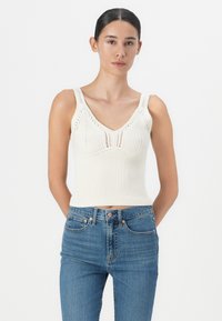 Hvit ribbet strikket tanktopp med V-hals og brede stropper, med intrikate åpen arbeidsdetaljer, kombinert med blå høy midje jeans.