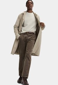 Beige Leinenmantel über einem weißen gerippten Pullover, kombiniert mit braunen Cordhosen und braunen Schnürschuhen. Das Model steht mit den Händen in den Taschen.