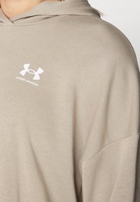 Sudadera con capucha color beige hecha de una tela suave, con un corte relajado y un pequeño logo blanco de Under Armour en el pecho.