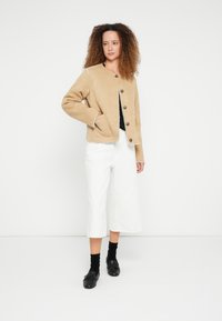 Beżowa zamszowa kurtka z okrągłym dekoltem i zapięciem na guziki, zestawiona z białymi spodniami culottes z wysokim stanem i czarnymi mokasynami. Teksturowane włosy.