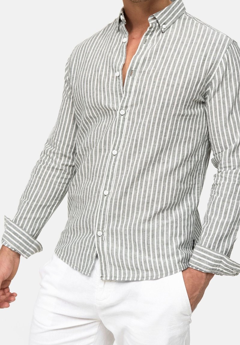 Man draagt een grijs-wit verticaal gestreept overhemd met opgerolde manchetten en witte broek, met een hand in de zak, tegen een effen achtergrond.