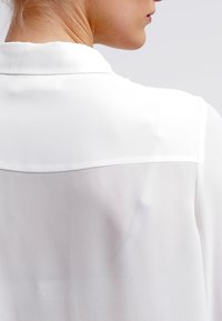 Blusa blanca, ligera, con una textura suave, que presenta un collar y un panel trasero transparente. Diseño simple sin patrones ni acentos adicionales.