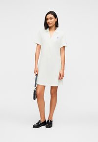 Φόρεμα σε στυλ πόλο από ανοιχτό πράσινο μείγμα βαμβακιού, με κοντά μανίκια, V-λαιμόκοψη, λεπτομέρεια με λογότυπο στο στήθος, φορεμένο με μαύρα μοκασίνια και μικρή τσάντα χειρός.