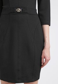 Robe noire ajustée à manches trois-quarts, ceinture décorative avec boucle circulaire dorée à la taille, montrée de la moitié de la cuisse au bras.