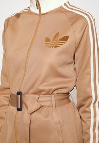 Hnědá sportovní bunda s vysokým límcem, zipem, bílými pruhy a opaskem v pase, na prsou s logem.