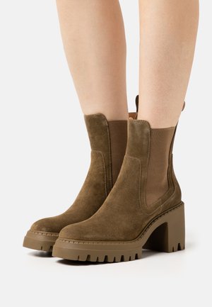 Brown suede ankle boots with chunky high heels and elastic side panels worn on bare legs against a white background.