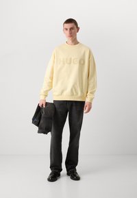 Sweatshirt amarelo claro com logotipo "HUGO" em relevo, mangas longas, punhos canelados, combinado com jeans escuros e segurando uma jaqueta preta dobrada.