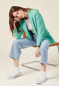 Blazer verde menta, camicia bianca, jeans larghi azzurri e sneakers bianche alte. Il modello è seduto su una sedia con sedile in legno.