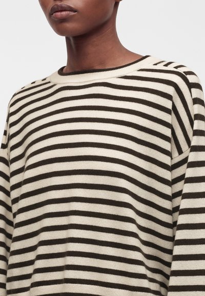Personne portant un pull en maille beige et noir à rayures horizontales avec un col rond, visible du cou au milieu du torse.