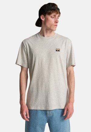 Giovane uomo con una t-shirt beige con grafica, jeans blu e un cappellino da baseball nero portato al contrario, che guarda verso sinistra.