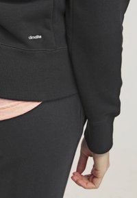 Sweatshirt noir avec des poignets côtelés, arborant un logo "climalite" discret. En dessous, un haut rose clair apparaît. Texture de tissu douce.