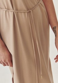 Beige šaty s přifasovaným pasem a šňůrkovým detailem, vyrobené z měkké látky, hladké textury a jemného lesku.