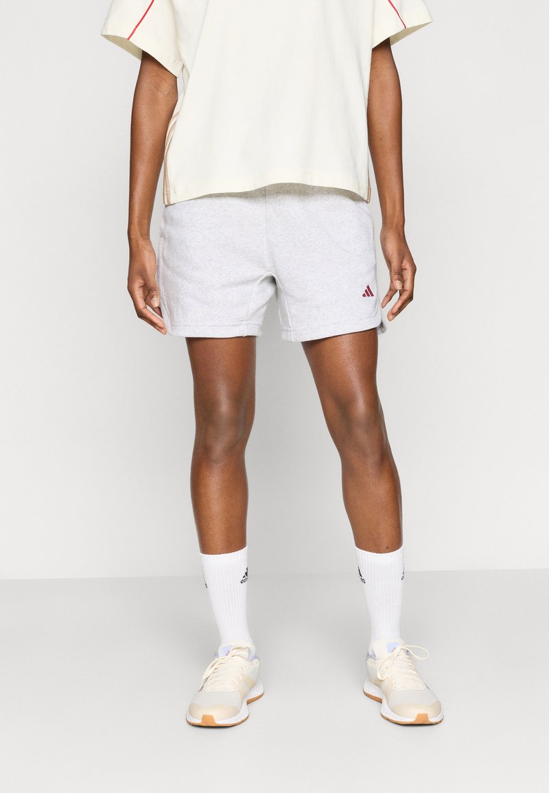 Sive bombažne kratke hlače z rdečim logotipom Adidas, kombinirane z belimi nogavicami in svetlimi športnimi copati, pod kremno oversized majico.