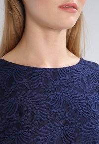 Námořnická modrá krajková halenka má květinový vzor, hladký povrch, lodičkový výstřih a krátké rukávy, které zdůrazňují jemné detaily.