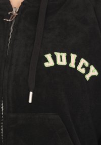 Felpa nera con zip in tessuto morbido, caratterizzata da una grande scritta ricamata "JUICY" in verde e rosa sul petto. Include tasche frontali e cordoncino.