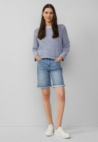 Suéter de punto azul claro con un patrón texturizado, combinado con shorts de mezclilla claros arremangados y zapatillas blancas. La modelo está de pie con las manos en los bolsillos.