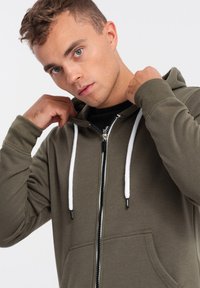 Olivově zelená mikina na zip vyrobená z měkké tkaniny, se bílými šňůrkami, předním zipem a dvěma bočními kapsami.