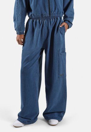 Persoon draagt loszittende blauwe broek met elastische tailleband en grote zijzak, gecombineerd met een bijpassende blauwe jas en witte sneakers.