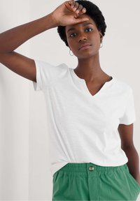 Fehér, rövid ujjú V-nyakú póló, puha textúrával, zöld, rugalmas derekú nadrággal, gombos díszítéssel párosítva.