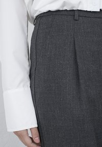 Grå skræddersyede bukser med et tekstureret stof, der har plisseringer og sidelommer. En hvid langærmet skjorte er synlig.