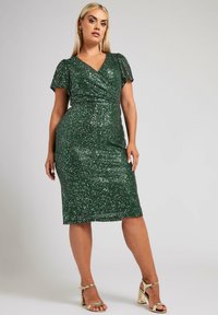 Vihreä paljettimekko, jossa on V-pääntie, lyhyet liehuvat hihat ja vartalonmyötäinen siluetti. Polvenmittainen, teksturoidulla kiiltopinnalla. Kultaiset korkokengät.