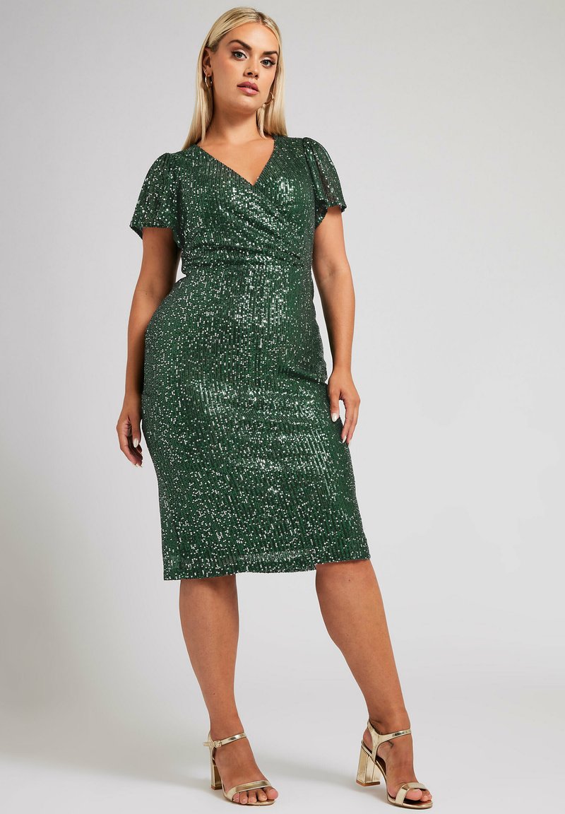 Vihreä paljettimekko, jossa on V-pääntie, lyhyet liehuvat hihat ja vartalonmyötäinen siluetti. Polvenmittainen, teksturoidulla kiiltopinnalla. Kultaiset korkokengät.