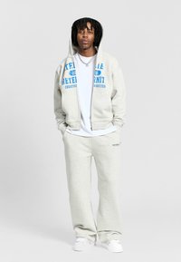 Szara bluza z kapturem z niebieskim nadrukiem, połączona z szarymi spodniami dresowymi. Materiał jest miękki, casualowy design z zamkiem z przodu i kieszeniami.