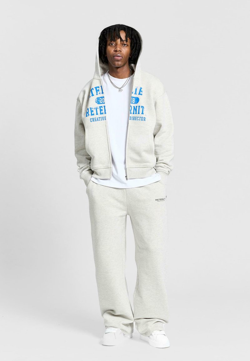 Szara bluza z kapturem z niebieskim nadrukiem, połączona z szarymi spodniami dresowymi. Materiał jest miękki, casualowy design z zamkiem z przodu i kieszeniami.