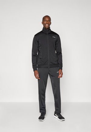 Mężczyzna stoi, ubrany w czarny dres Adidas z kurtką na zamek i dopasowanymi spodniami, czarne trampki z białymi podeszwami, uśmiechając się.