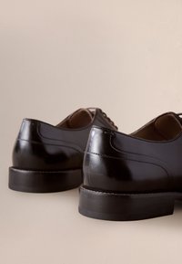 Paar polierte schwarze Lederschuhe, von hinten auf einer neutralen beigen Oberfläche gezeigt.