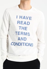 Weißes Sweatshirt mit langen Ärmeln und blauer Schrift, die "ICH HABE DIE ALLGEMEINEN GESCHÄFTSBEDINGUNGEN GELESEN" lautet. Es hat einen runden Halsausschnitt und gerippte Bündchen.