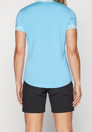 Personne portant une chemise bleu clair à manches courtes avec des poignets à motifs et un short noir, vue de dos sur un fond uni.