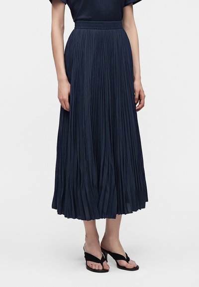 Femme portant une jupe midi plissée bleu marine taille haute et des sandales noires à talons bas sur un fond uni.