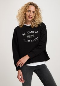 Czarna bluza z białym graficznym napisem. Okrągły dekolt, luźny krój, długie rękawy, noszona na białej koszuli. Miękka tkanina, minimalistyczny design.