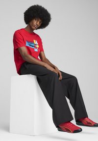 Rød t-shirt med et blåt grafisk design, parret med løse sorte bukser og røde sko med sorte og gule accenter, siddende på en hvid blok.