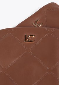 Hnědé kožené doplňky mají embosovaný vzor s textem "ILOLA CASADEMUNT." Kovové logo "LC" dodává výjimečný detail.