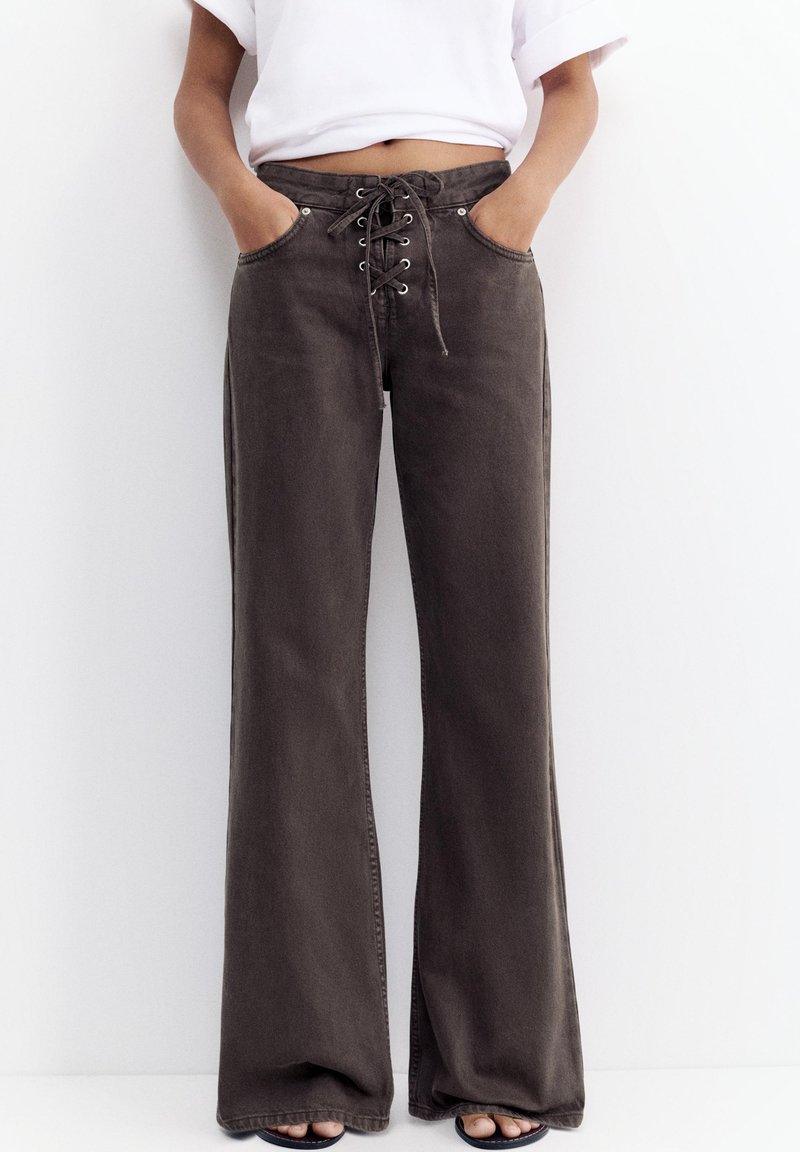 Personne portant un jean noir à jambes larges avec laçage à l'avant et mains dans les poches, associé à un t-shirt blanc court et des sandales noires.