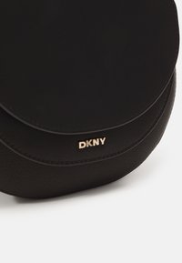 Borsa a tracolla nera in pelle con patta arrotondata e logo DKNY dorato sul fronte, esposta su uno sfondo bianco.