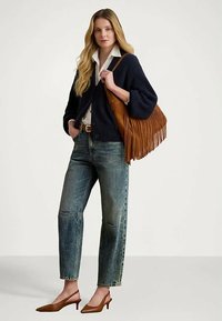 Mulher vestindo um cardigan azul marinho, camisa branca, jeans azuis, sapatos de salto slingback castanhos, segurando uma bolsa de ombro com franjas castanha.