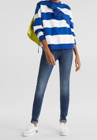 White and blue striped sweater, casual fit, paired with dark blue skinny jeans and white sneakers with blue accents, accessorized with a green bag.