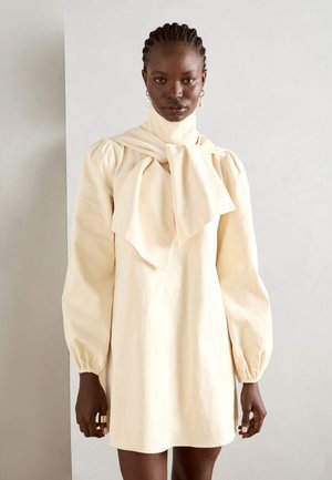 Cream-colored, long-sleeve dress with a high neckline and large bow accent. Made from textured fabric, featuring puffed sleeves and a relaxed fit.