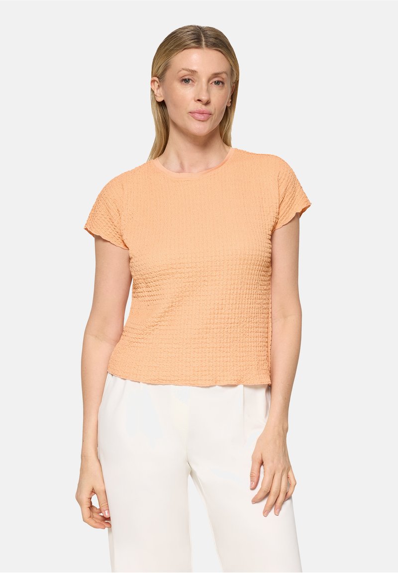 Femme aux cheveux blonds raides portant un haut pêche texturé à manches courtes et un pantalon blanc, debout devant un fond clair uni.